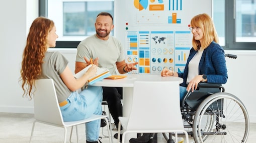A - Drei Personen im Gespräch an einem runden Tisch in einem modernen Büro; eine Person nutzt einen Rollstuhl.