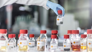 A hand with a blue glove holds a small bottle with a red lid over a row of similar bottles on a table in the laboratory.