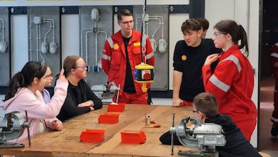 Jugendliche sitzen an einem Werkbanktisch und sprechen mit Auszubildenden in roter Schutzkleidung von Shell während einer praktischen Übung am Girls’ und Boys’ Day.