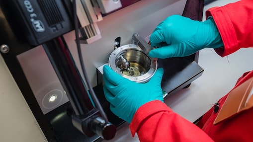 Hände in blauen Handschuhen, die an einem metallischen Gerät in einer Laborumgebung arbeiten.