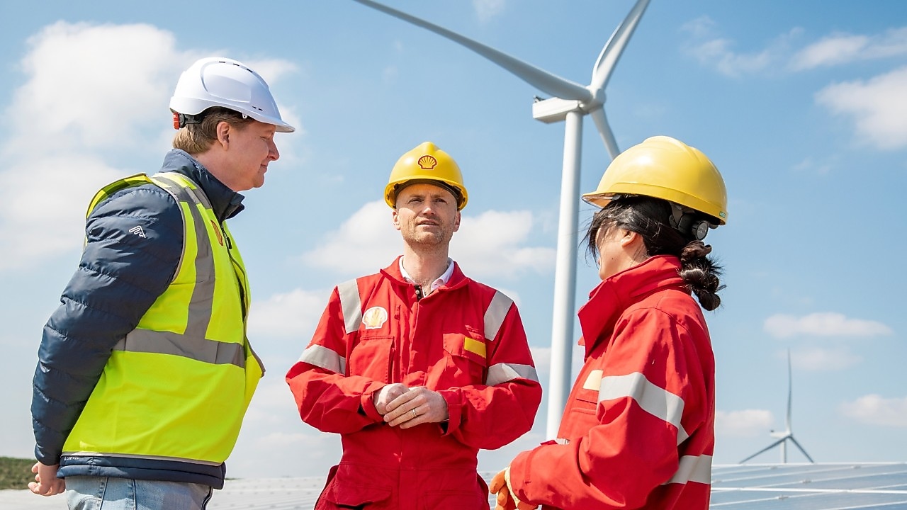 Drei Fachkräfte in Sicherheitskleidung stehen draußen bei Windkraftanlagen und diskutieren.