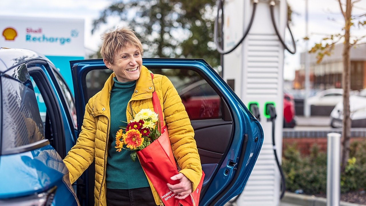 Eine Frau steigt aus einem blauen Elektroauto und hält eine Tasche mit Blumen in der Hand. Sie lächelt, als sie an einer Shell Recharge-Lades