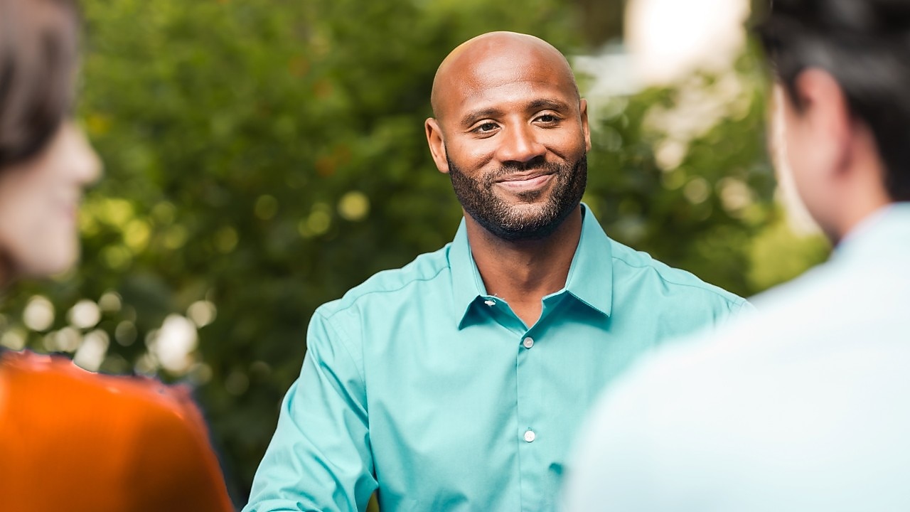 Ein Mann lächelt, während er mit zwei Personen spricht, die teilweise im Bild zu sehen sind.