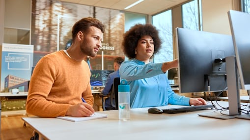 Person zeigt auf einen Computermonitor, während eine andere Person mitschreibt