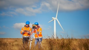 Zwei Ingenieure mit blauen Helmen und orangefarbenen Westen begutachten ein Feld mit Geräten und einer Windturbine im Hintergrund