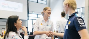 Zwei Personen schütteln sich in einem geschäftlichen Umfeld die Hände und lächeln sich freundlich zu, während eine dritte Person zusieht.