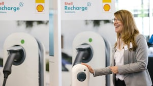 Eine Frau im Geschäftsanzug lächelt neben einem Shell Recharge Ladegerät für Elektrofahrzeuge.