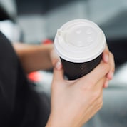Hände halten einen Kaffeebecher aus braunem Papier mit einem weißen Deckel.