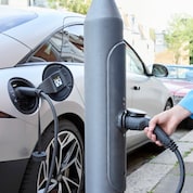 Eine Hand schließt ein Stromkabel an eine Ladestation neben einem Auto auf einer Straße an.