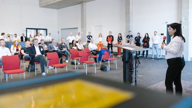 Sechs Personen stehen an weißen Podien vor einem hellblauen Hintergrund und lächeln selbstbewusst.