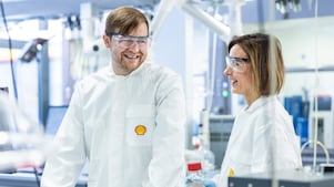 Zwei Labortechniker in weißen Kitteln und Schutzbrillen unterhalten sich in einem Labor.
