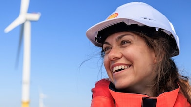 Eine lächelnde Frau mit Schutzhelm und Schwimmweste steht im Freien am Meer vor Windkraftanlagen.