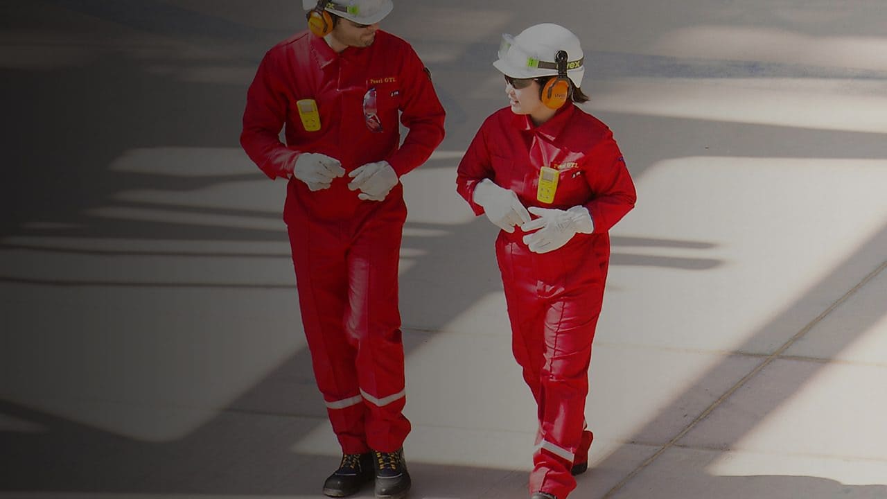 Zwei Arbeiter in roten Shell-Uniformen und Helmen gehen nebeneinander auf einem Industriegelände.