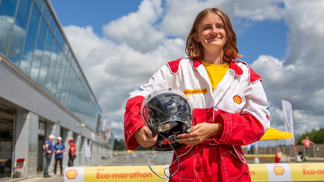 Junge Person in einem rot-weißen Rennanzug hält lächelnd einen Helm.