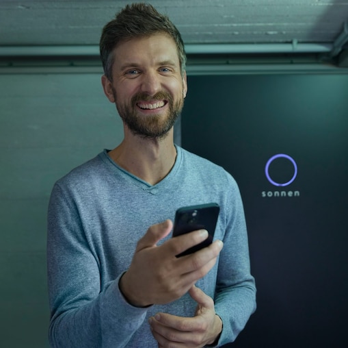 Lächelnder Mann mir einem Smartphone vor einer sonnen Batterie