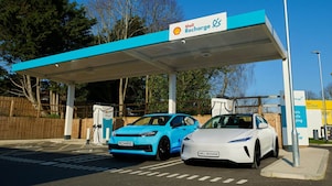 Zwei Elektroautos (blau und weiß) laden unter einem überdachten Shell Recharge-Schnellladepunkt. Im Hintergrund ein Holzzaun, Bäume und klarer Himmel an einem sonnigen Tag.