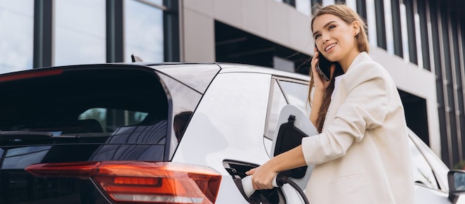Junge Frau telefoniert lächelnd während sie ihr E-Auto lädt.