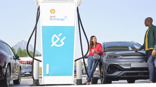 Junge Frau unterhält sich mit einem jungen Mann, während  sie ihr E-Auto an einer Shell Recharge Ultraschnellladesäule lädt.