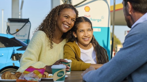 Eine Frau und ein Kind sitzen lächelnd an einem Tisch im Freien an einer Shell Recharge Ladestation. Ein Mann sitzt ihnen gegenüber. Im Hintergrund ist ein Elektrofahrzeug an einer Ladesäule zu sehen.
