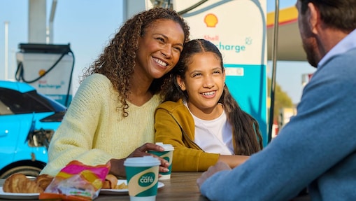 Eine Frau und ein Kind sitzen lächelnd an einem Tisch im Freien an einer Shell Recharge Ladestation. Ein Mann sitzt ihnen gegenüber. Im Hintergrund ist ein Elektrofahrzeug an einer Ladesäule zu sehen.