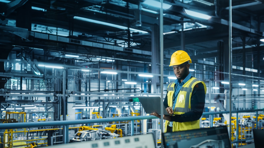 Ein männlicher Ingenieur, der einen gelben Helm und eine Warnweste trägt, steht mit einem Laptop in einer modernen, hoch technisierten Autofabrik.