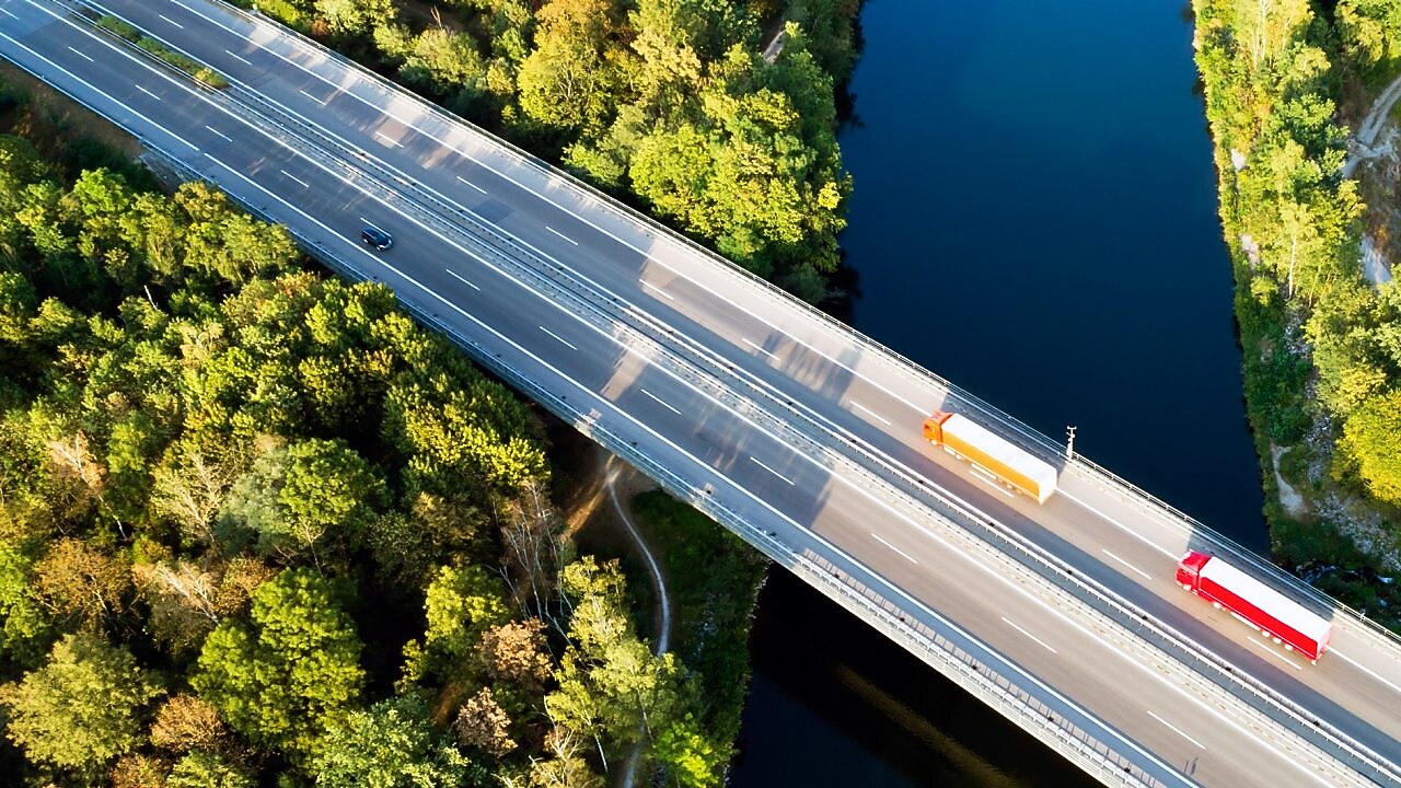 Aus der Vogelperspektive führt eine Brücke in einen grünen Mischwald. Darauf fahren ein gelber und roter Lkw sowie entgegenkommend ein Auto.