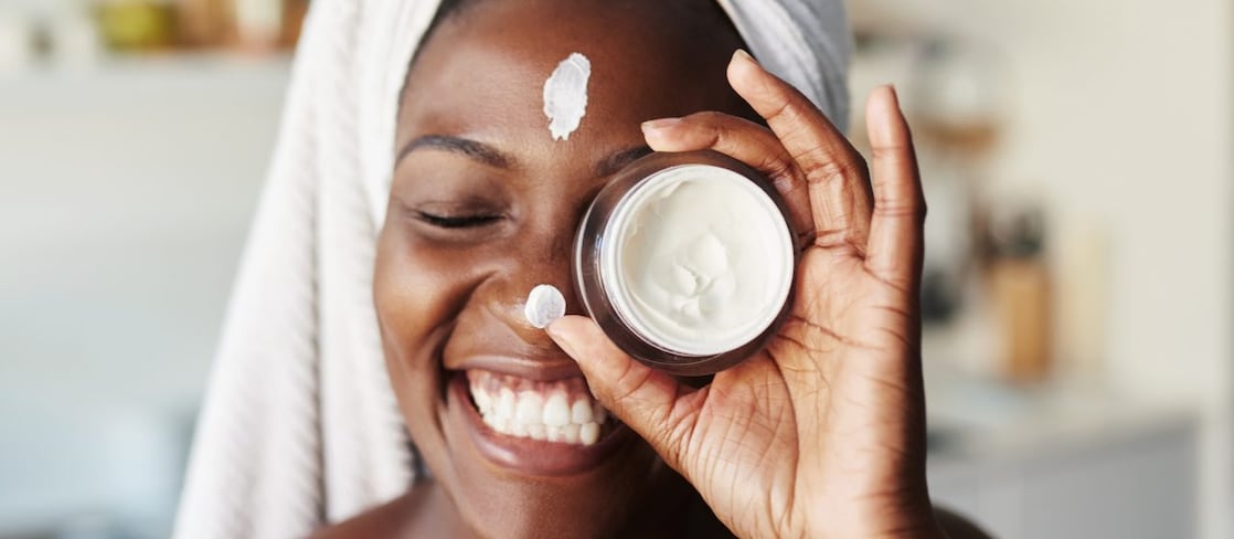 Eine Person mit einem um den Kopf gewickelten Handtuch hält ein Objekt vor ihr Gesicht. Im Hintergrund sind Regale mit verschiedenen Gegenständen zu sehen, was auf ein Badezimmer oder eine Küche hindeutet. Das Gesicht der Person ist unscharf. 