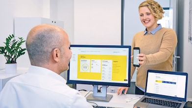 Eine Frau steht vor einem Mann, der an einem Schreibtisch sitzt und zeigt ihm über den Tisch ein Smartphone. Auf dem Schreibtisch stehen ein Computer und ein Laptop – auf beiden ist Shell Fleet Hub geöffnet.