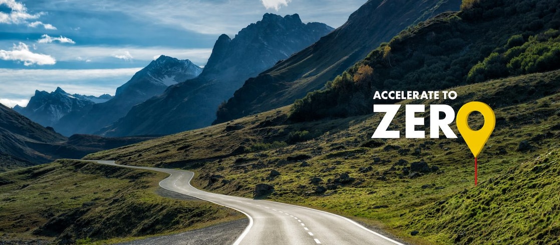 Auto fährt auf einer Schnellstraße durch grüne Landschaft mit Bergen im Hintergrund, Textaufschrift: "Accelerate to Zero".