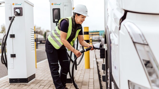 Eine Lkw-Fahrerin mit Warnweste und weißem Helm steht an einem Shell Recharge Depot und steckt gerade den Ladestecker in den E-Lkw.