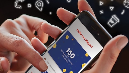 Nahaufnahme einer Frauenhand, welche ein Handy mit geöffneter Shell App in der Hand hält. Um das Handy herum fliegen Icons zu Autowäsche, Mehrwegbecher, Rabatten, Zapfpistole.