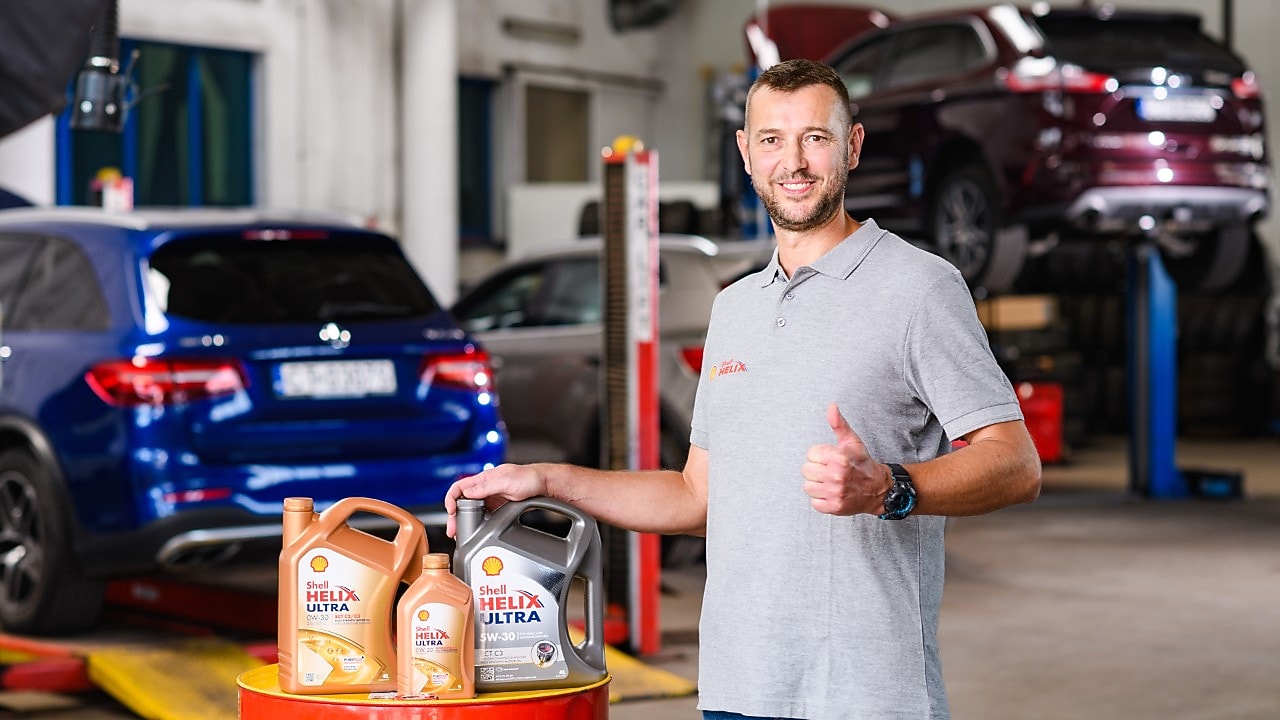 Kfz‑Mechaniker in moderner Autowerkstatt präsentiert Shell Helix Ultra Motoröle (0W‑30, 5W‑30) vor angehobenen Fahrzeugen – hochwertige Motoröle für optimalen Motorschutz und Leistung