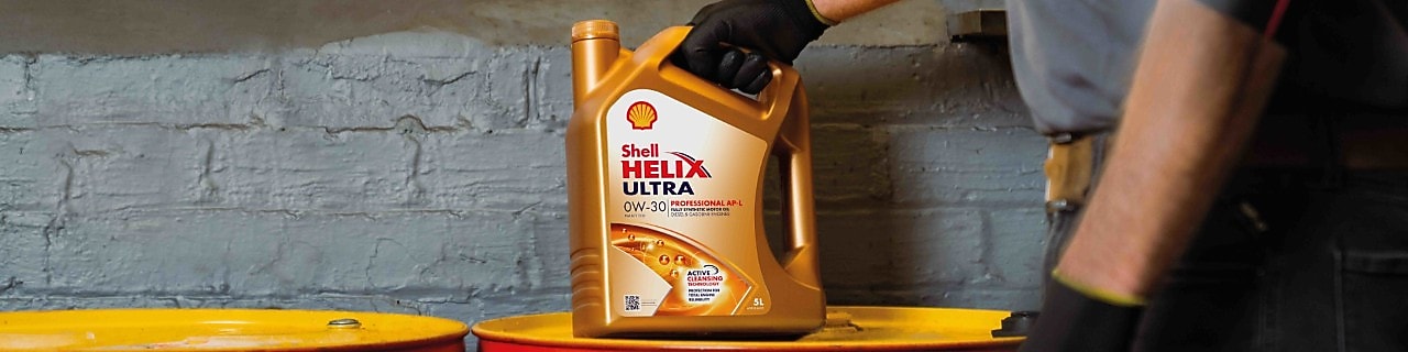 Mechaniker hält einen Kanister Shell Helix Ultra 0W‑30 Motoröl über gelben Ölfässern in einer Werkstatt.