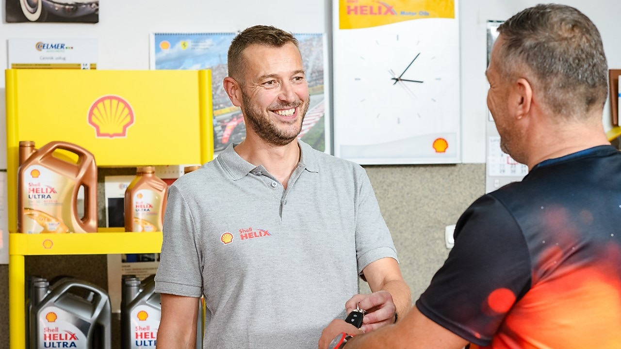 Servicepersonal in einer Werkstatt übergibt ein Shell‑Helix‑Produkt an einen Kunden; im Hintergrund Shell‑Helix‑Ultra‑Motoröl auf einem Regal und Shell‑Markenmaterialien.