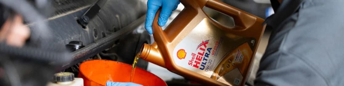 „Mechaniker mit blauen Schutzhandschuhen gießt Shell Helix Ultra 0W‑30 Motoröl aus einem goldenen Kanister in einen orangefarbenen Trichter, um frisches Motoröl in den Motor eines Fahrzeugs einzufüllen.“