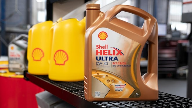 „Shell Helix Ultra 0W‑30 Motorölkanister steht im Vordergrund auf einer Werkbank, flankiert von gelben Shell‑Behältern in einer professionellen Autowerkstatt; Produktetikett und Markenlogo sind klar sichtbar.“
