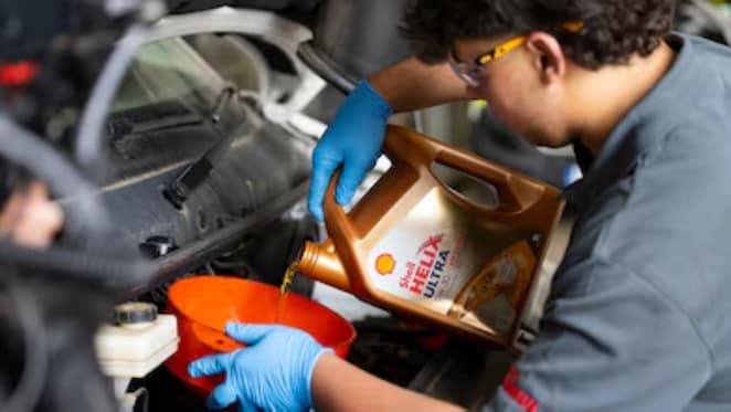 „Mechaniker mit blauen Schutzhandschuhen gießt Shell Helix Ultra 0W‑30 Motoröl aus einem goldenen Kanister in einen orangefarbenen Trichter, um frisches Motoröl in den Motor eines Fahrzeugs einzufüllen.“