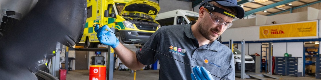 Werkstattmitarbeitender überprüft mit einem Ölmessstab den Ölstand eines Fahrzeugs, während im Hintergrund mehrere gelbe Einsatzfahrzeuge mit geöffneten Motorhauben sowie Shell Helix Werbematerialien in einer professionellen Servicehalle zu sehen sind.