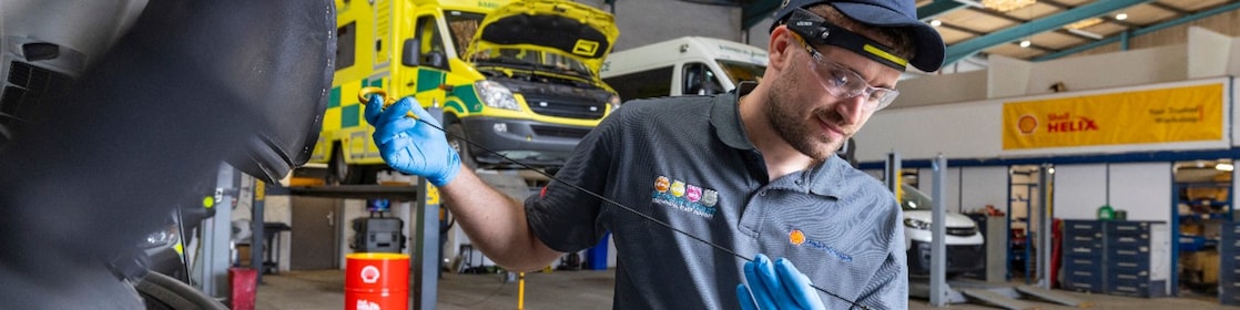 Werkstattmitarbeitender überprüft mit einem Ölmessstab den Ölstand eines Fahrzeugs, während im Hintergrund mehrere gelbe Einsatzfahrzeuge mit geöffneten Motorhauben sowie Shell Helix Werbematerialien in einer professionellen Servicehalle zu sehen sind.