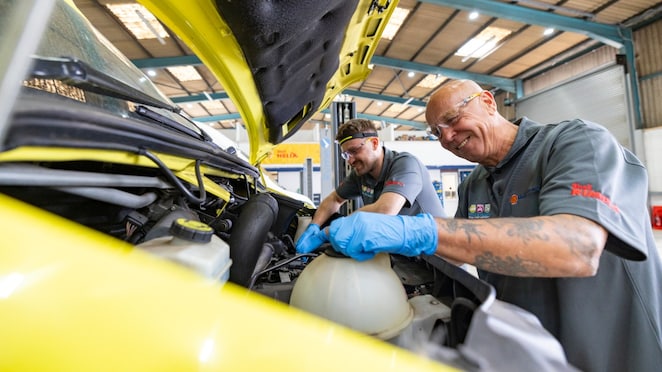 Zwei Werkstattmitarbeitende arbeiten im Motorraum eines gelben Fahrzeugs, während die Motorhaube geöffnet ist; einer nutzt blaue Schutzhandschuhe und führt Wartungsarbeiten im vorderen Bereich des Motors aus.