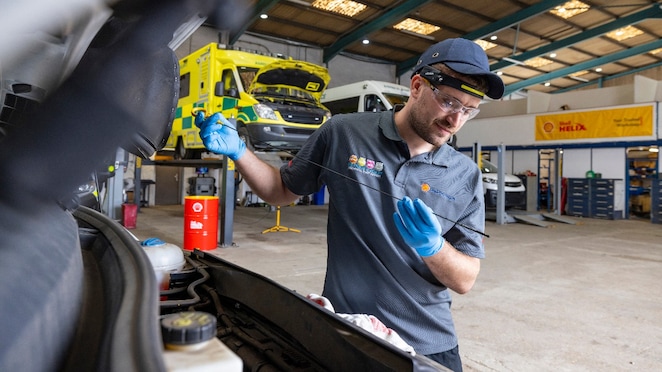 Werkstattmitarbeitender überprüft mit einem Ölmessstab den Ölstand eines Fahrzeugs, während im Hintergrund mehrere gelbe Einsatzfahrzeuge mit geöffneten Motorhauben sowie Shell Helix Werbematerialien in einer professionellen Servicehalle zu sehen sind.