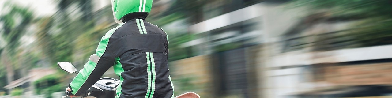 Motorradfahrer mit grünem Helm und schwarzer Jacke mit grünen Streifen fährt auf einem Roller durch eine städtische Umgebung, dargestellt in dynamischer Bewegung.