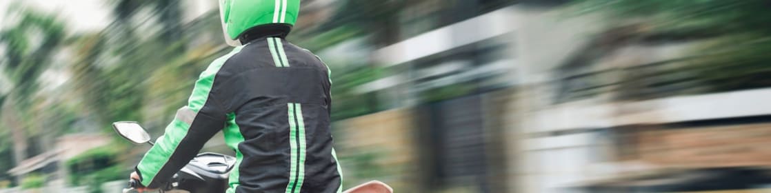 Motorradfahrer mit grünem Helm und schwarzer Jacke mit grünen Streifen fährt auf einem Roller durch eine städtische Umgebung, dargestellt in dynamischer Bewegung.