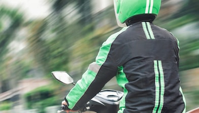 Motorradfahrer mit grünem Helm und schwarzer Jacke mit grünen Streifen fährt auf einem Roller durch eine städtische Umgebung, dargestellt in dynamischer Bewegung.