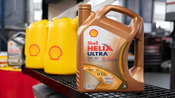 Motorölkanister Shell Helix Ultra 0W‑30 auf einem Werkstattregal, im Hintergrund stehen mehrere gelbe Shell‑Kanister in einer Kfz‑Werkstatt.