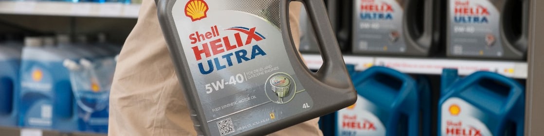 Person hält einen 4‑Liter‑Kanister Shell Helix Ultra 5W‑40 Motorenöl vor einem Regal mit weiteren Shell‑Motorenölen in einem Geschäft; Produktetikett und Markenlogo sind klar sichtbar.