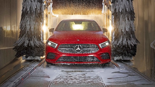 Eine rote Mercedes-Benz Limousine fährt durch eine Shell Car Wash Waschanlage. Das Fahrzeug wird beidseitig von großen rotierenden Bürsten umgeben, Wassertropfen und Schaum sind sichtbar. Die Scheinwerfer des Fahrzeugs sind eingeschaltet, der Mercedes-Stern im Kühlergrill ist deutlich erkennbar.