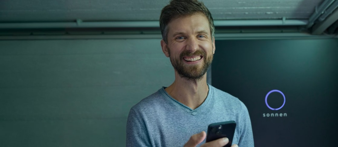 Lächelnder Mann mir einem Smartphone vor einer sonnen Batterie
