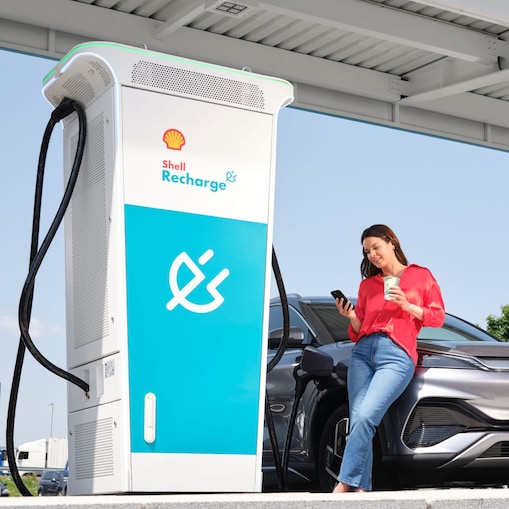 Frau lehnt an ihrem E-Auto und trinkt Kaffee, während das Fahrzeug an einer Shell Recharge Ultraschnellladesäule lädt.