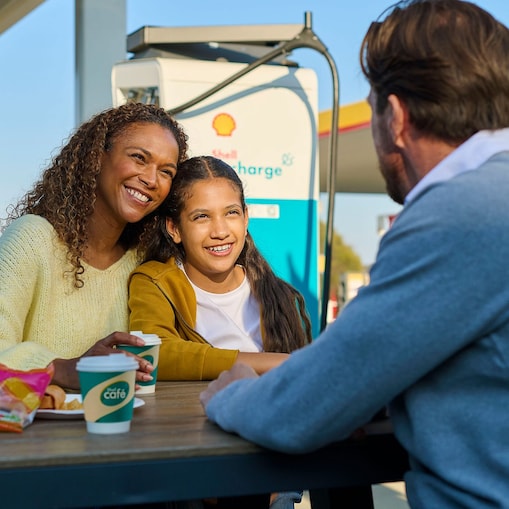 Eine Frau und ein Kind sitzen lächelnd an einem Tisch im Freien an einer Shell Recharge Ladestation. Ein Mann sitzt ihnen gegenüber. Im Hintergrund ist ein Elektrofahrzeug an einer Ladesäule zu sehen.using charge card at a Shell Recharge Station
